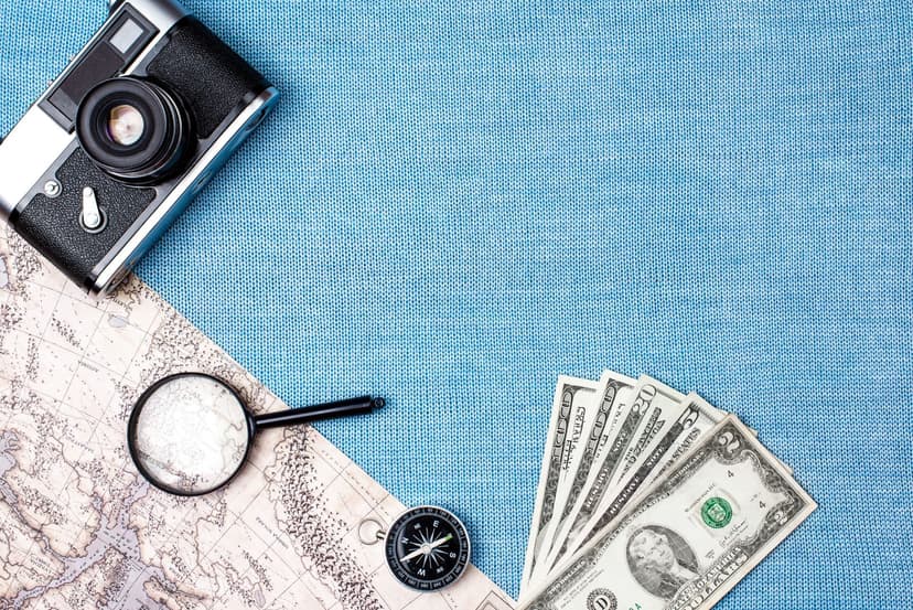 Money, a compass, and a camera laid out on the ground on top of a blue blanket and some sand