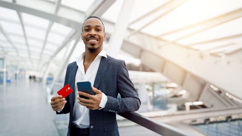 A man holding his phone and a credit card, smiling in an airport.